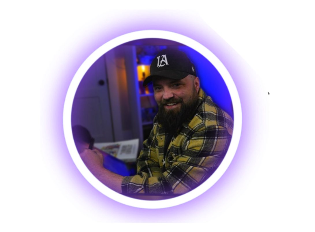 A man with a beard wearing a black baseball cap and a yellow plaid shirt, smiling at the camera, sitting indoors at night, illuminated by blue and pink lighting.