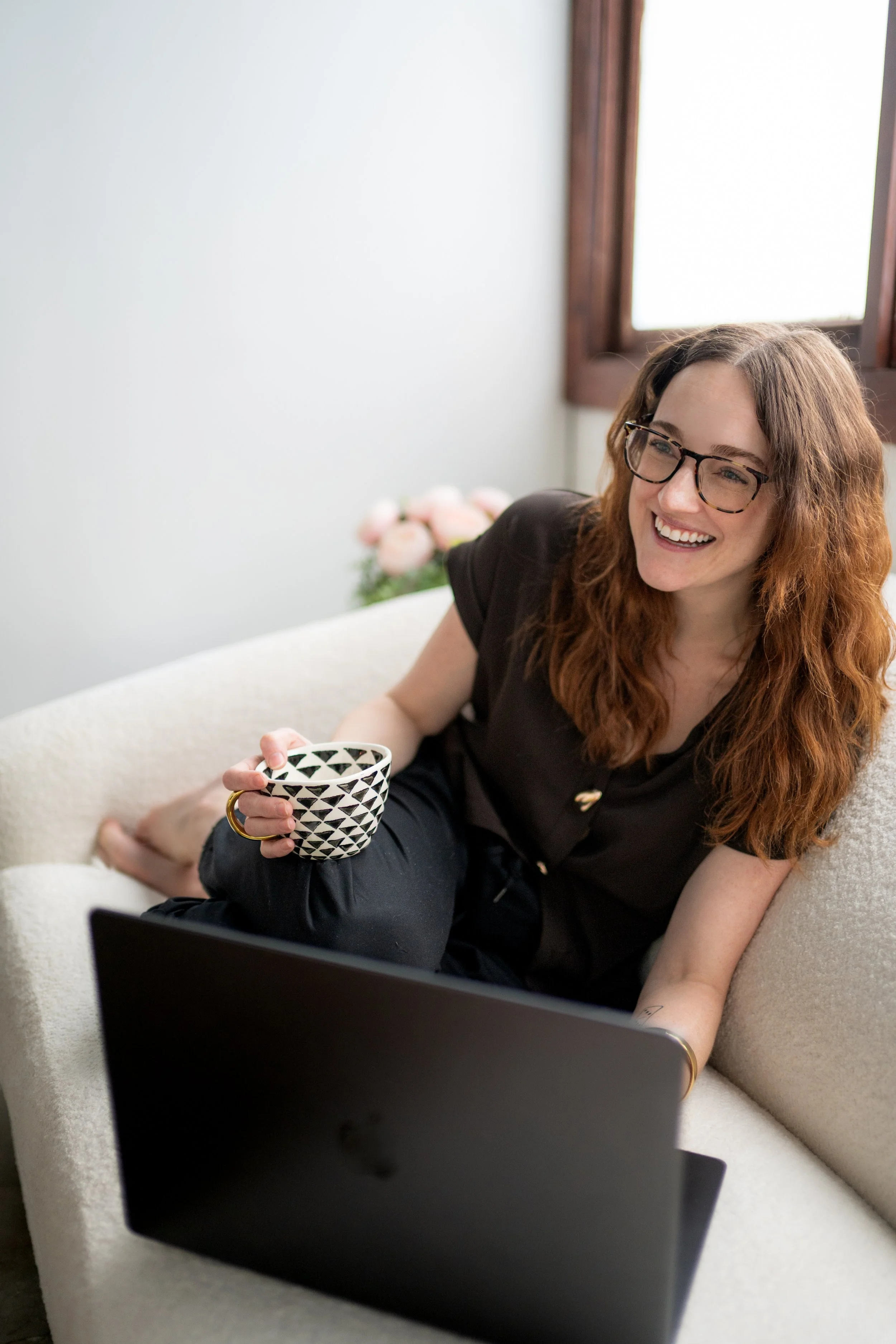 A woman with red hair and glasses sitting on a white sofa, smiling, holding a black and white patterned mug and working on a laptop, with a bouquet of pink flowers in the background.
