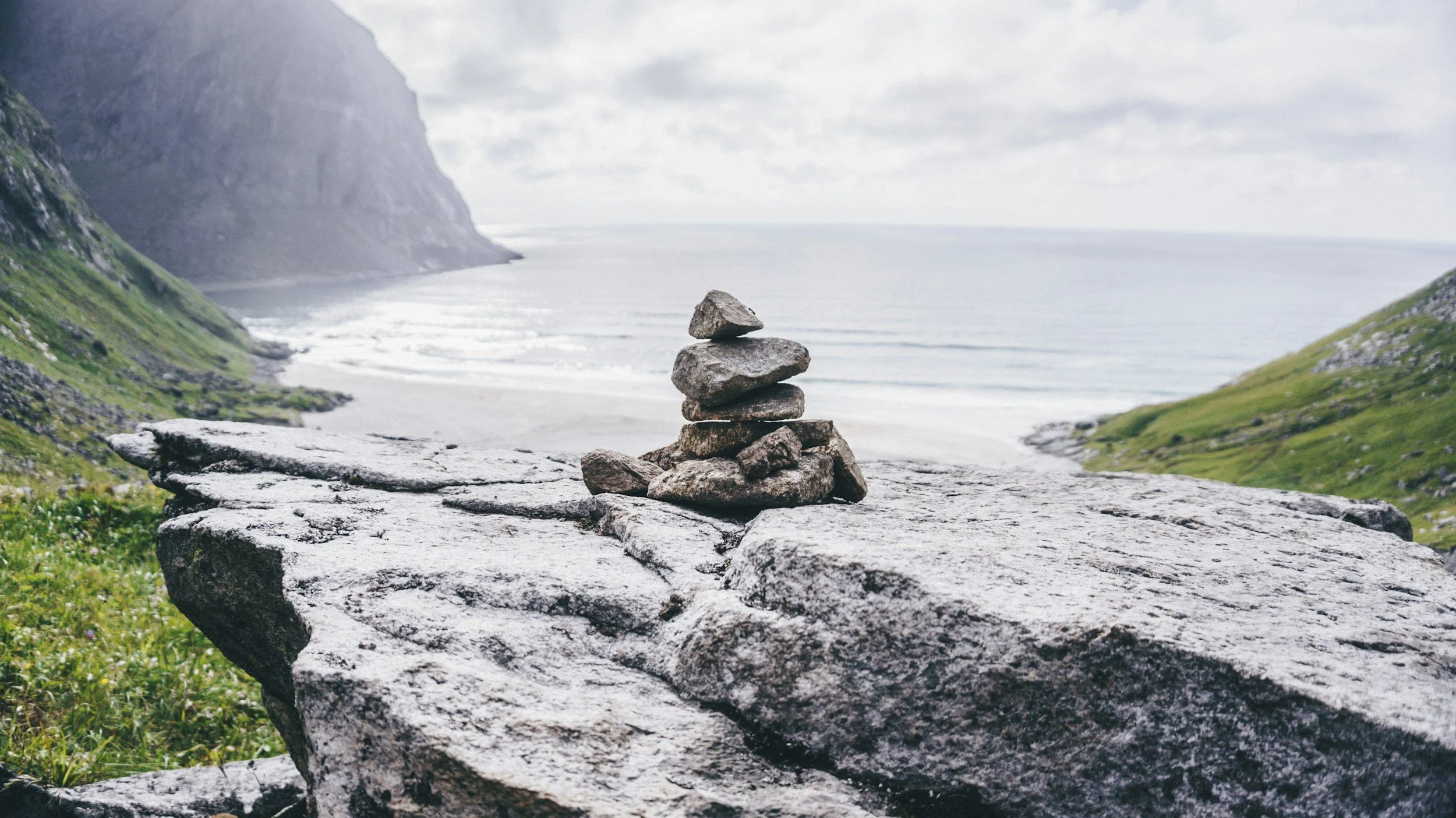 A stack of rocks balanced on a large flat stone on a hillside overlooking the ocean with cliffs on either side and cloudy sky above.