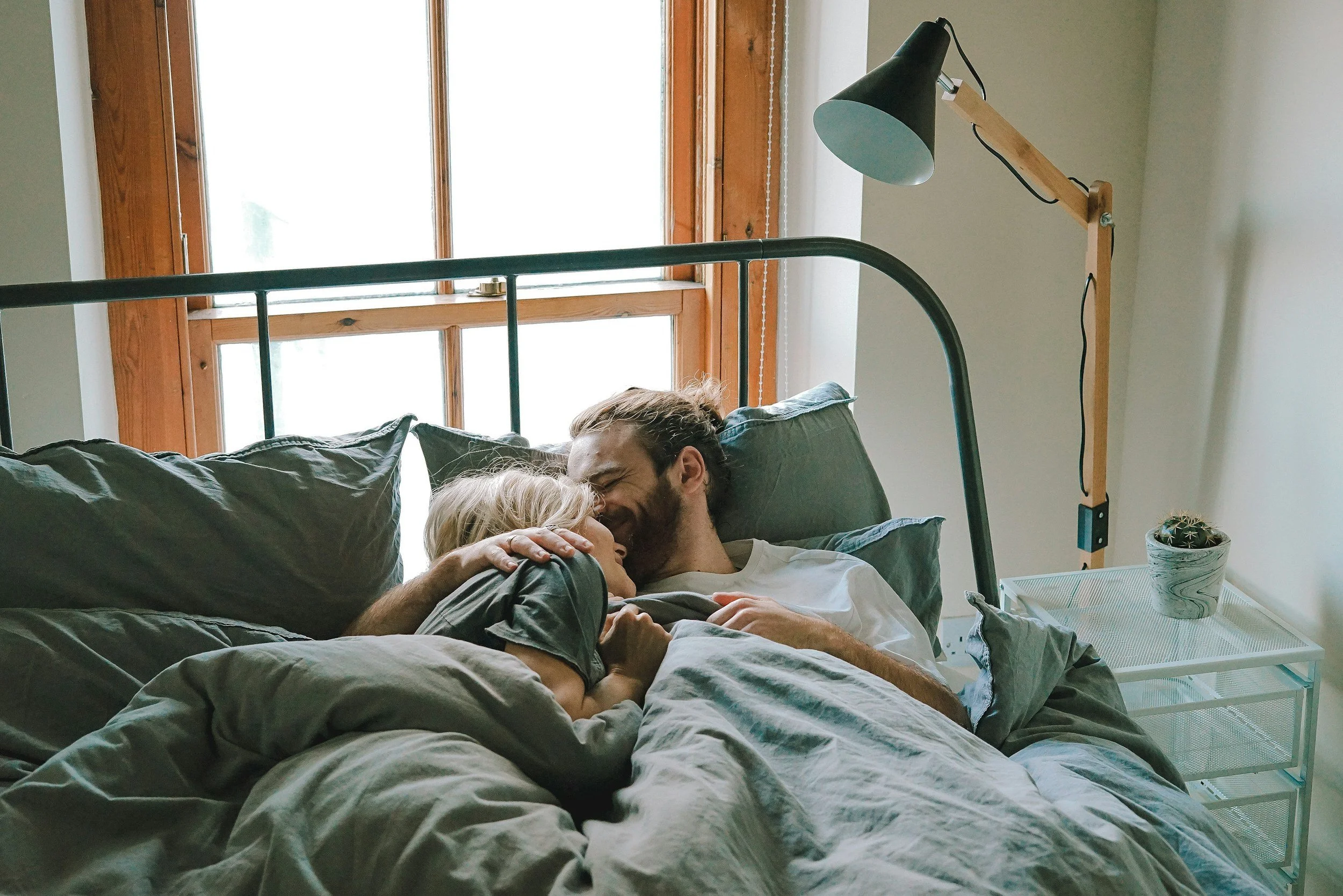 A man and a woman sharing a kiss while lying in bed, with a bedside table and a cactus plant beside them, and a large window in the background.
