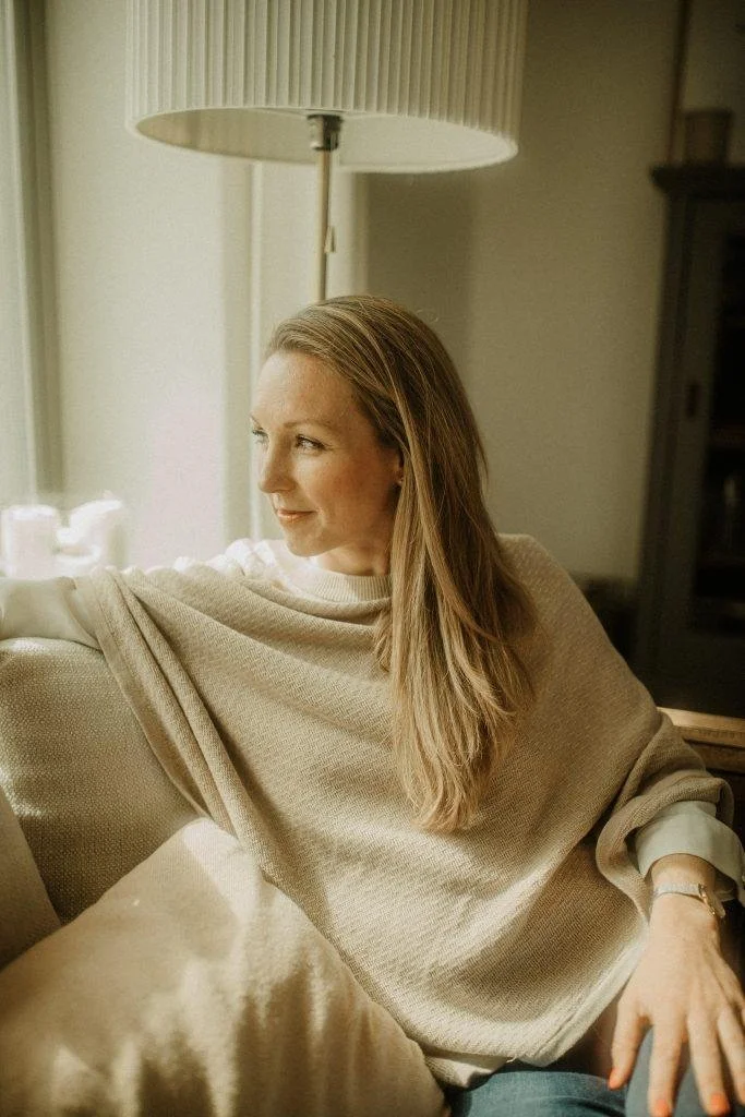 Woman sitting on a couch by a window with sunlight, wearing a beige sweater, looking out thoughtfully.