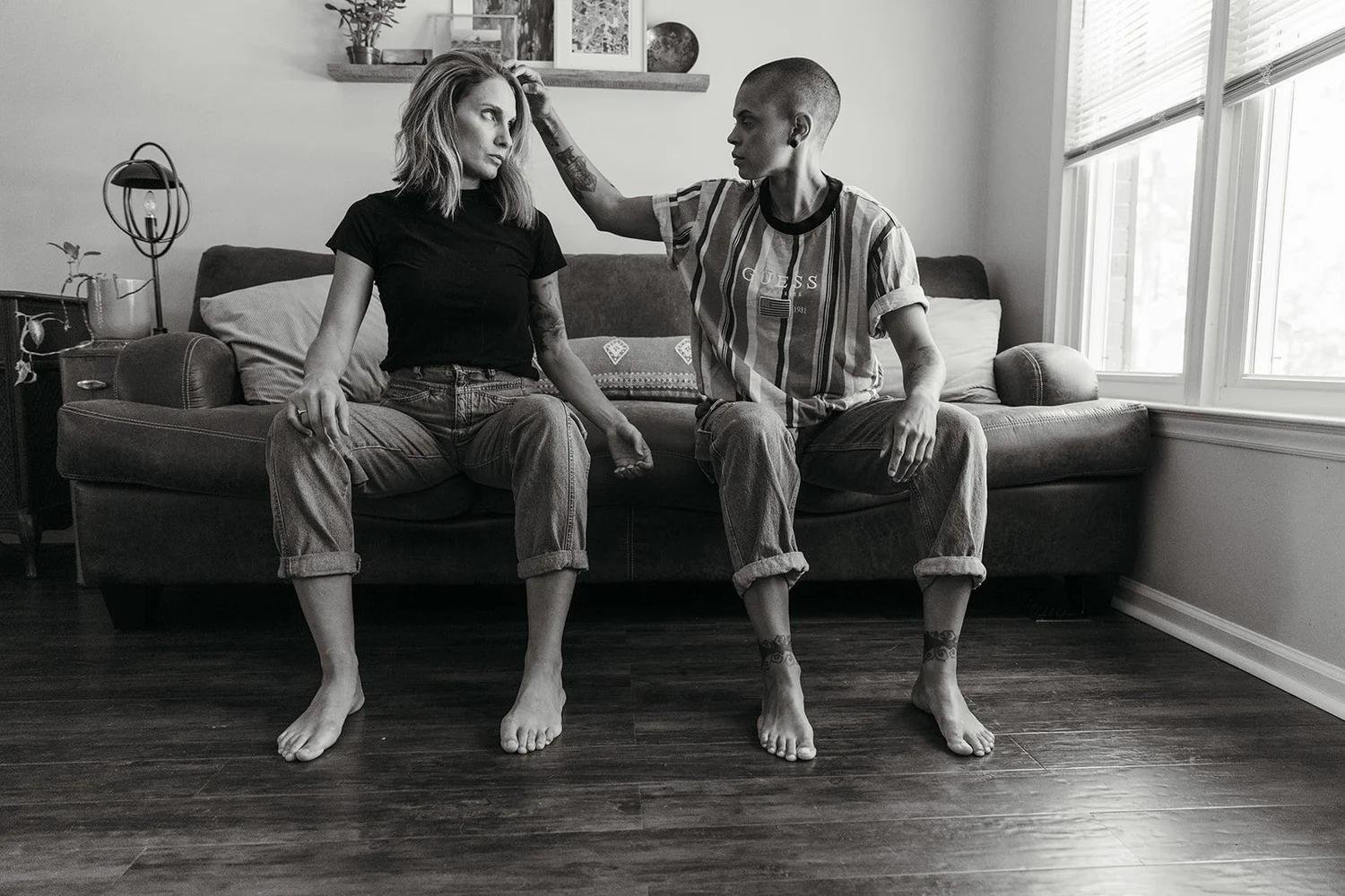Black and white photo of two young women sitting on a couch in a living room. One woman has shaved head and tattoos, wearing a striped T-shirt, touching the other woman's head. The woman with long hair is wearing a black T-shirt and jeans. Both are barefoot, and the scene is lit by natural light from large windows.
