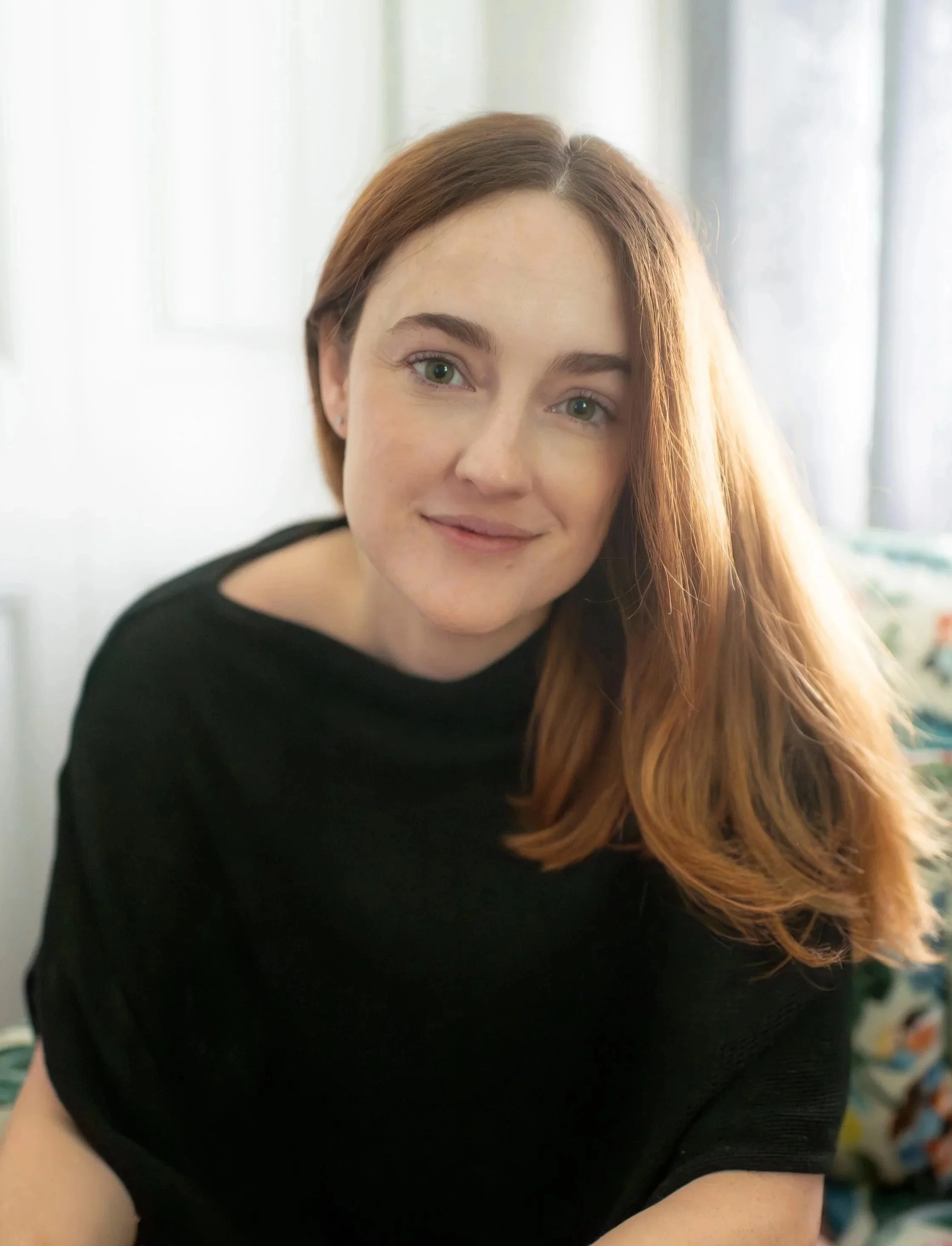 A woman with long red hair and blue eyes wearing a black top, smiling in a softly lit room with curtains in the background.