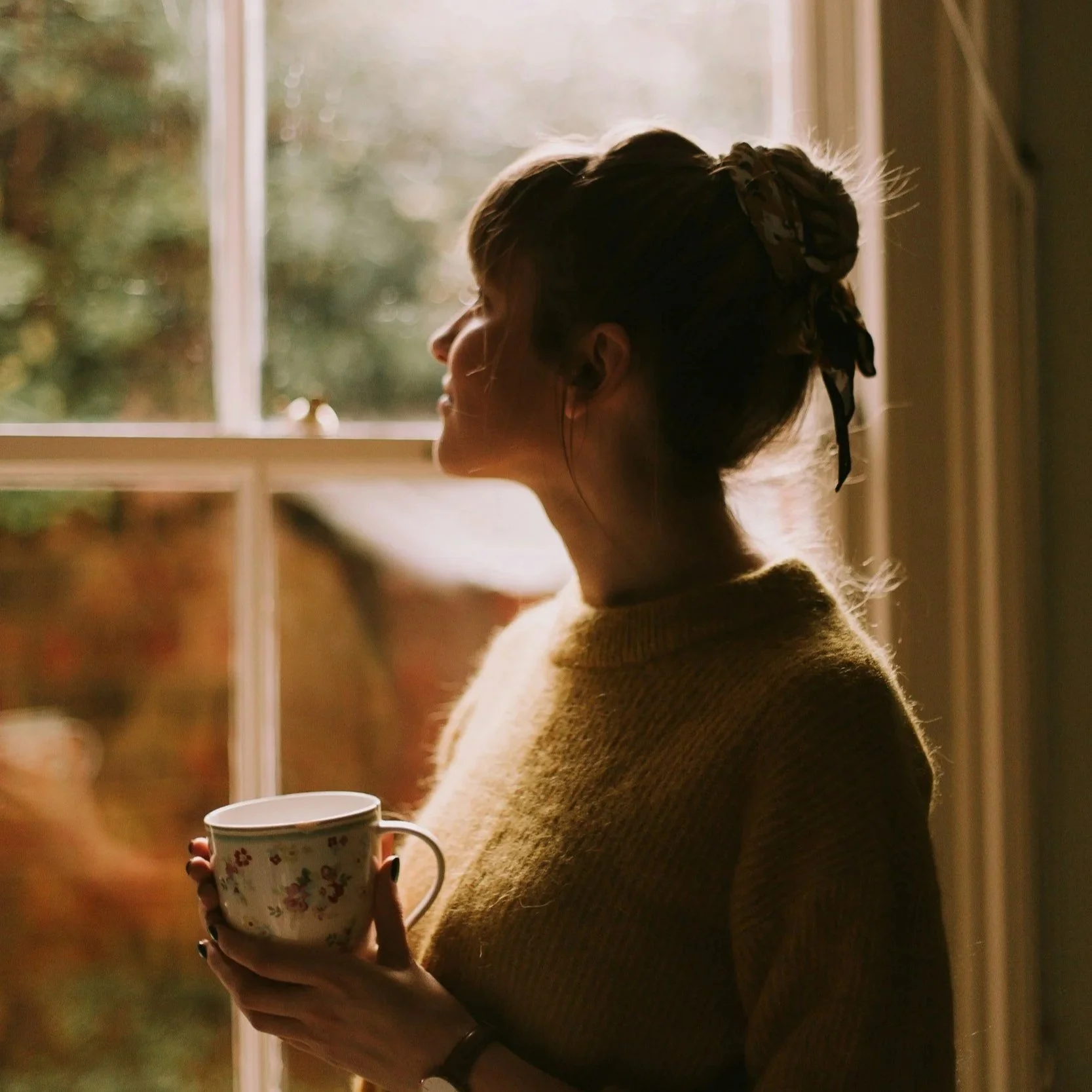 A woman standing in front of a window holding a mug, with sunlight shining through and illuminating her face and hair.