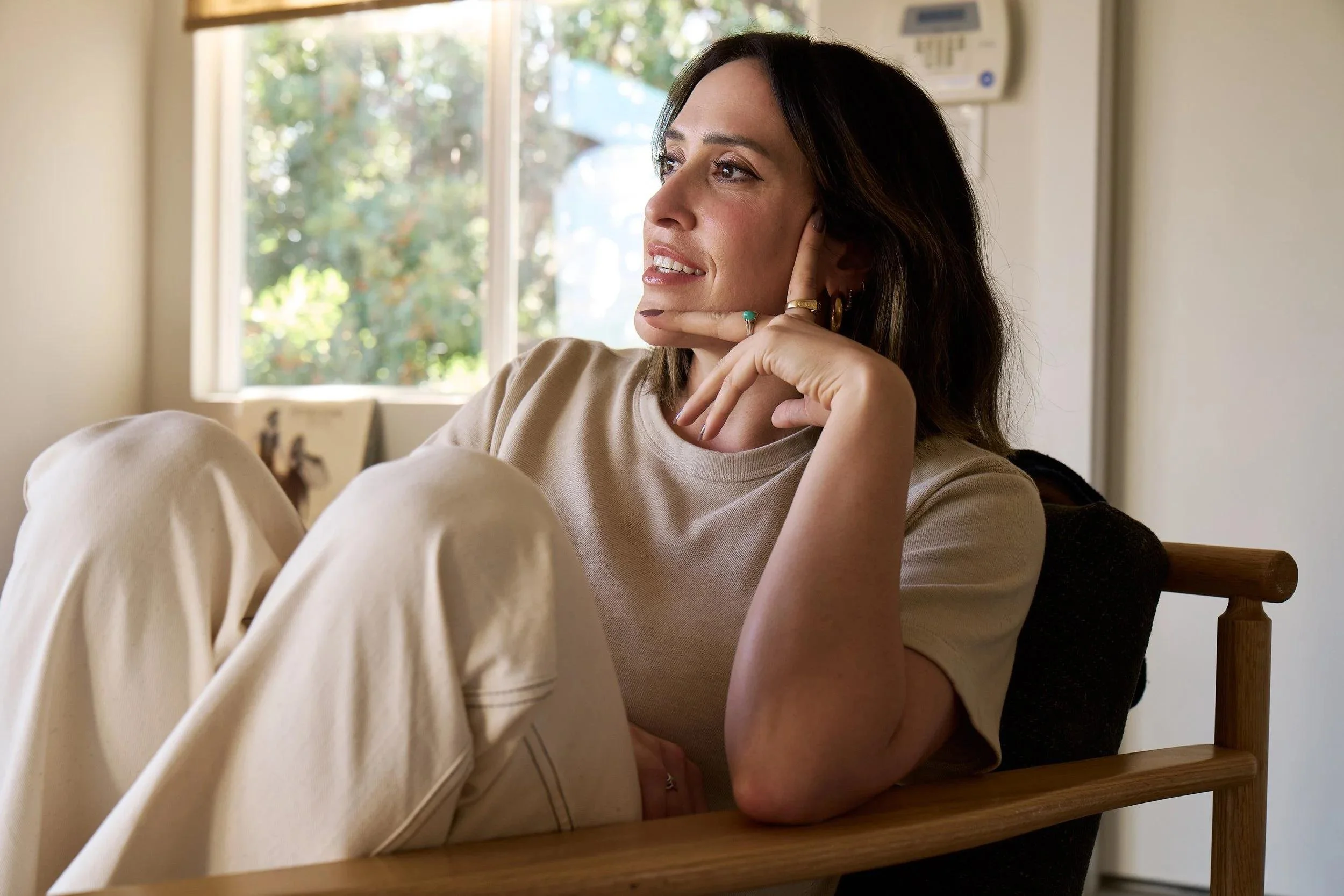 A woman with dark hair and wearing rings rests her chin on her hand, sitting on a wooden chair near a window with greenery outside. She has a calm, thoughtful expression.