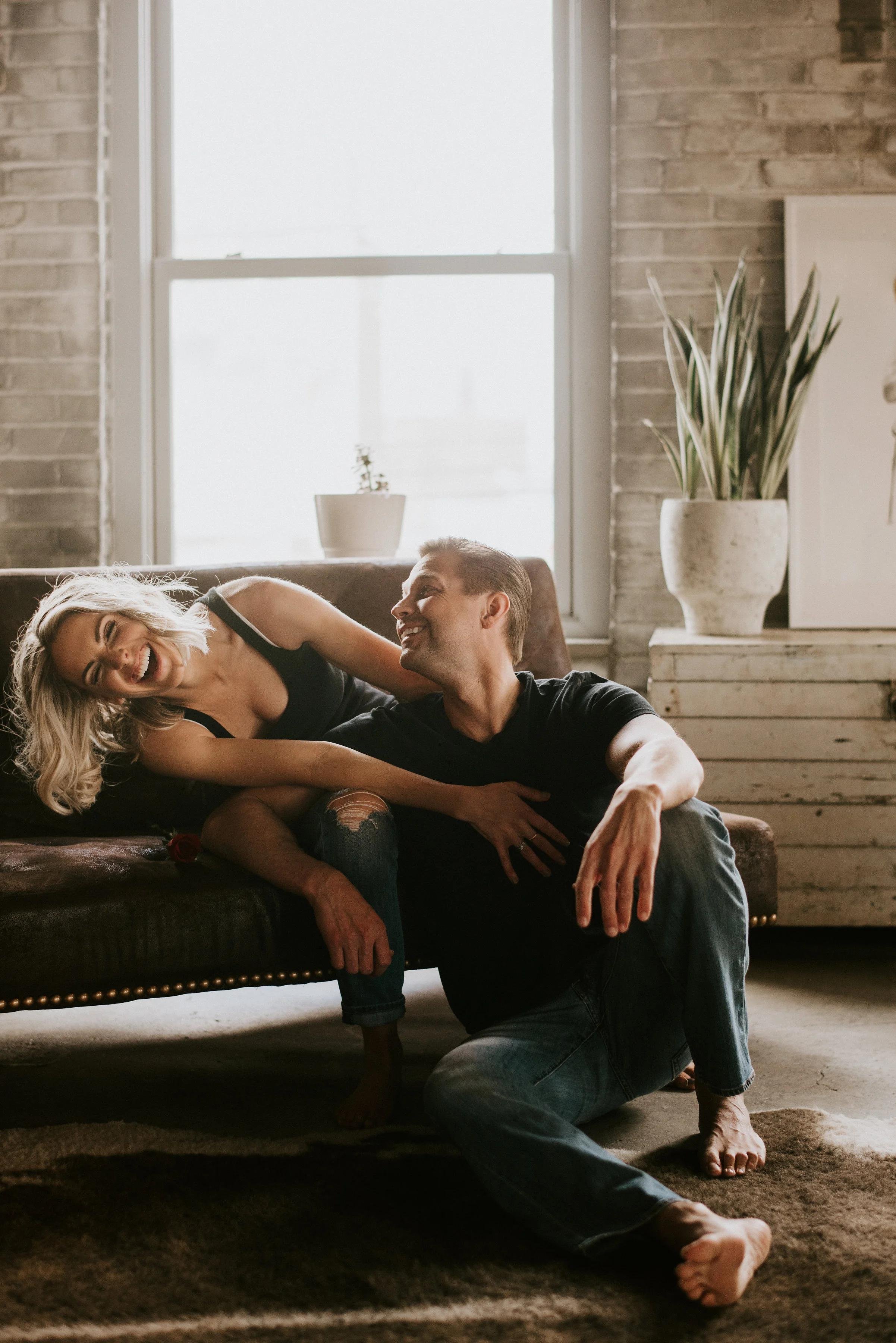 A happy couple sitting on a vintage-style sofa in a cozy, rustic apartment with a large window, houseplants, and a brick wall in the background.
