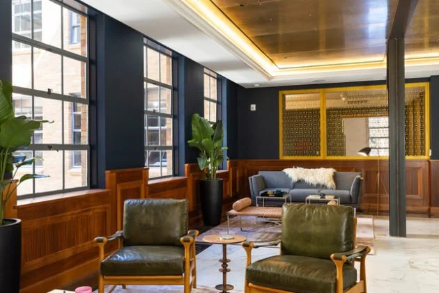 Modern lounge with large windows, wooden wainscoting, green leather chairs, a gray sofa with a white throw, and potted plants.