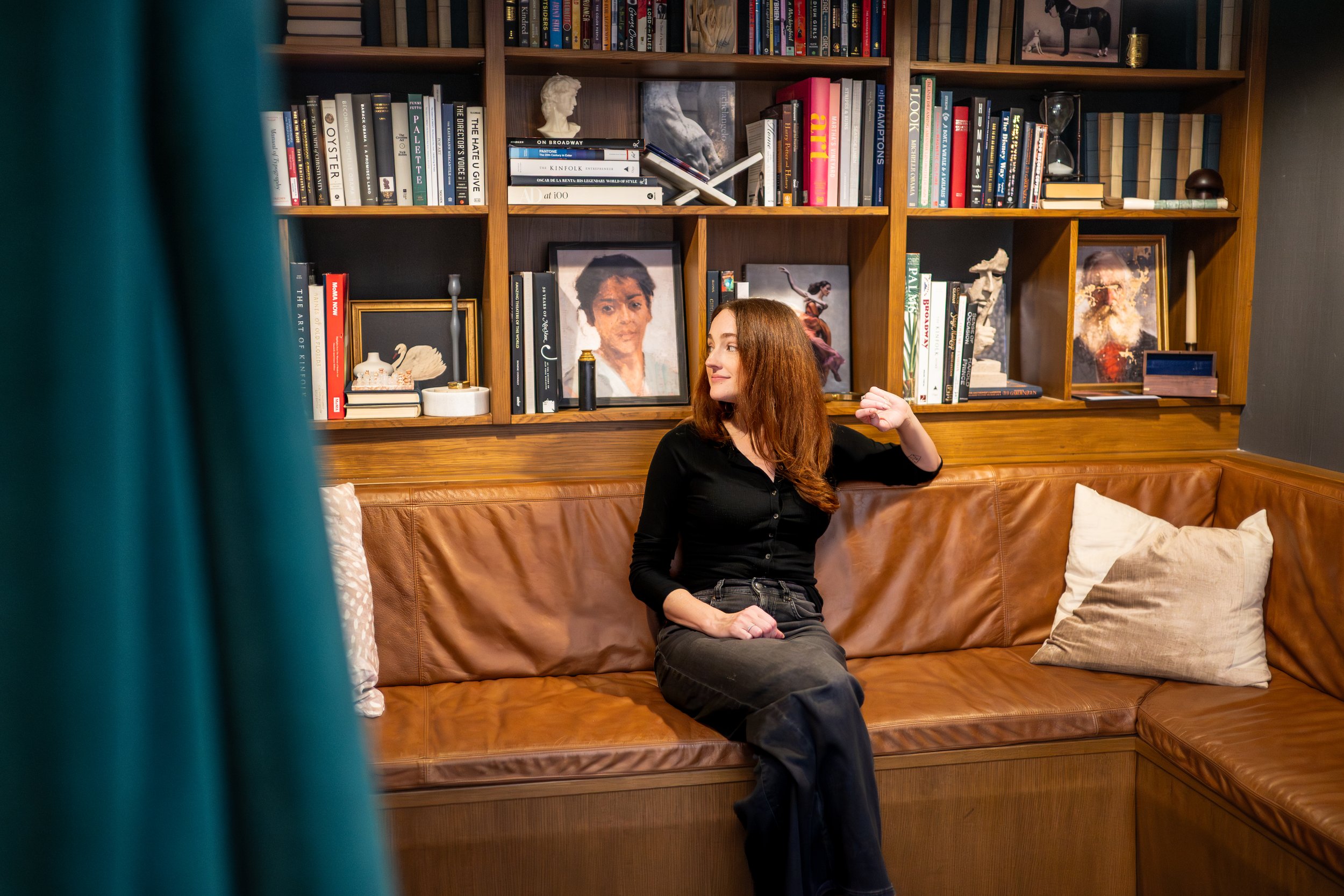A woman with long red hair, wearing a black top and dark pants, sitting on a brown leather couch in a room with a wooden bookshelf. The woman is looking to her left and smiling, with a small tattoo visible on her left wrist.