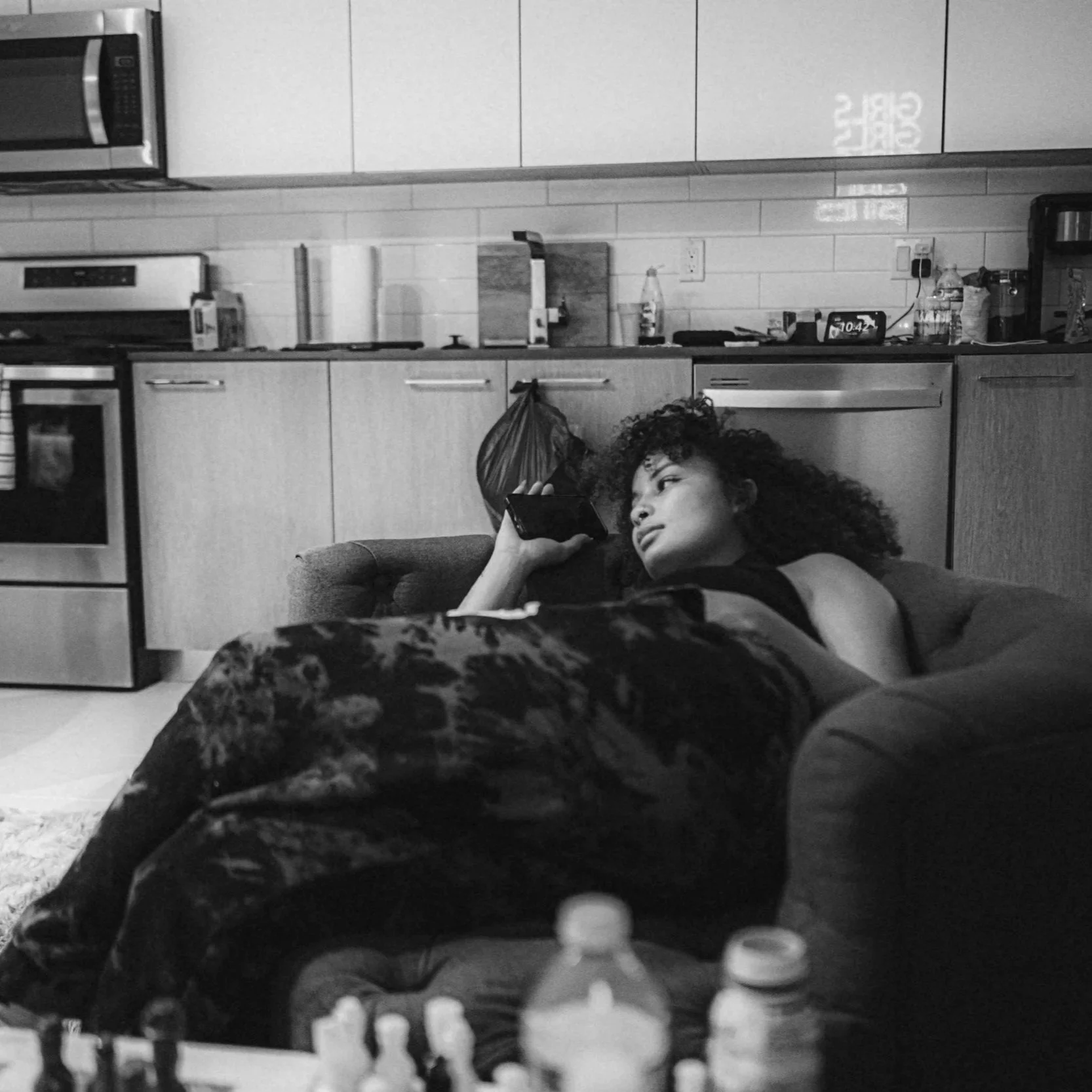 A woman with curly hair lying on a couch in a kitchen, holding a phone, with various bottles and kitchen appliances on the counter behind her.