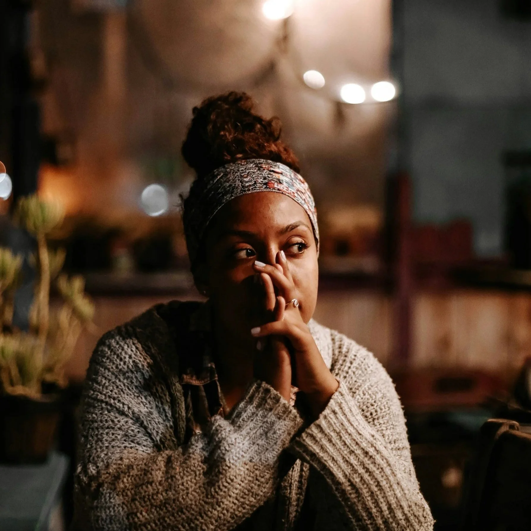 A woman with a patterned headscarf covering her hair, wearing a knitted sweater, is in a dimly lit setting with blurred lights in the background. She is touching her face with one hand and gazing thoughtfully to the side.