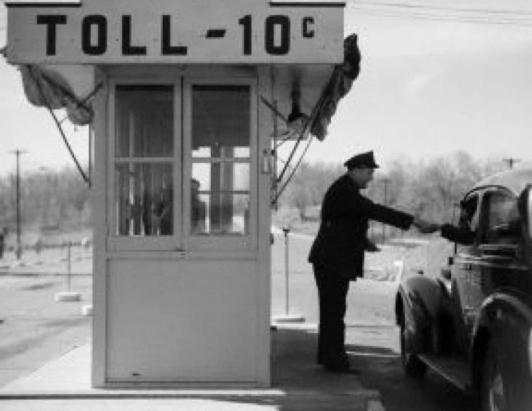 Driver handing coins to a toll collector at a mid-20th-century highway toll booth