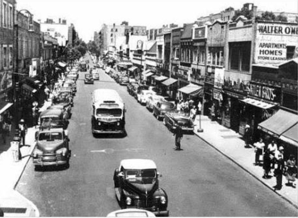 Street view of Queens in the 1950s showing apartment buildings, shops, and everyday neighborhood life.