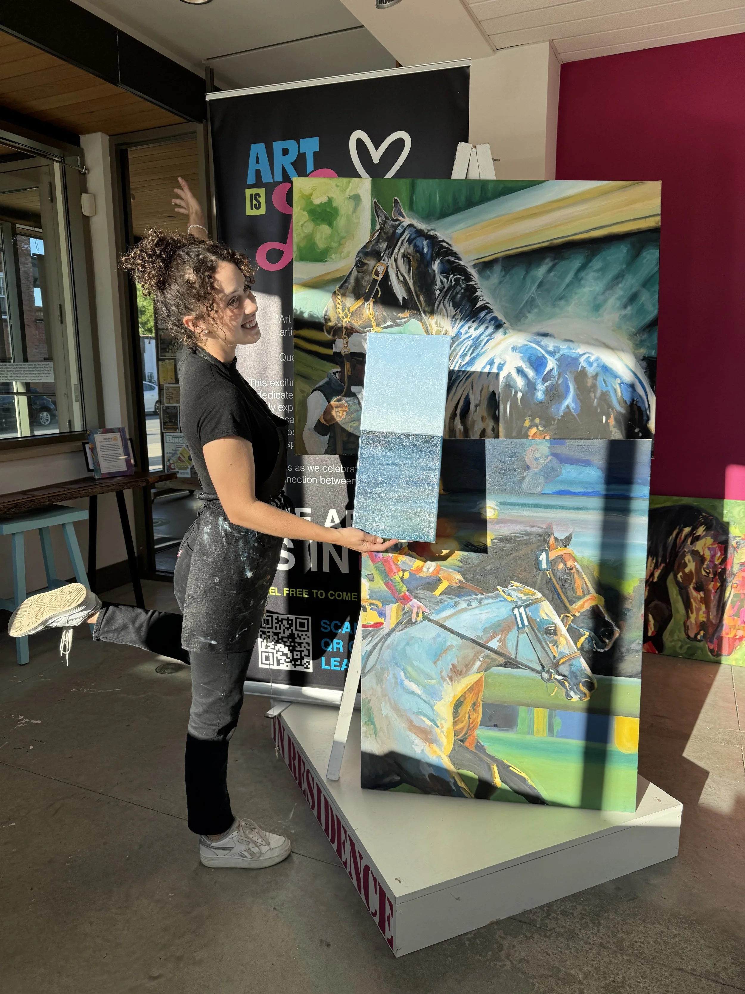 A woman with curly hair, wearing a black shirt and paint-stained pants, is smiling and holding a small painting of a beach scene with water and sky. She stands next to large, colorful paintings of racehorses and a horse in a stable, with a background advertisement banner promoting art events and a QR code.