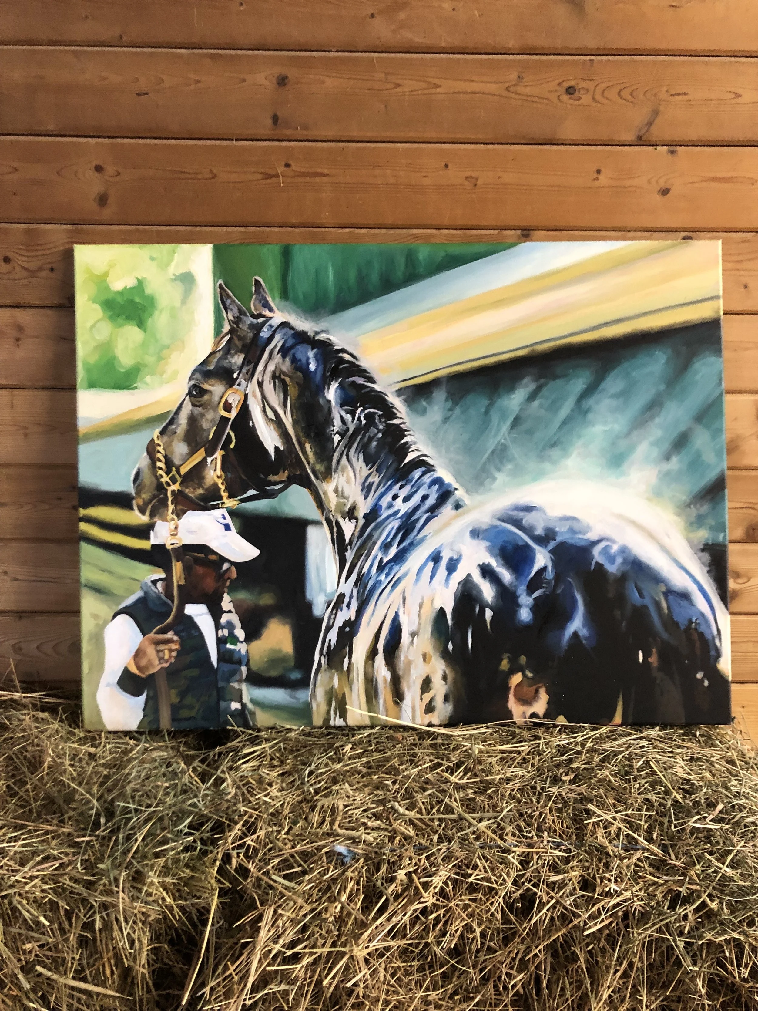 Painting of a horse with a person holding a lead rope in front, set against a green and blue background, displayed on a hay bale against a wooden wall.