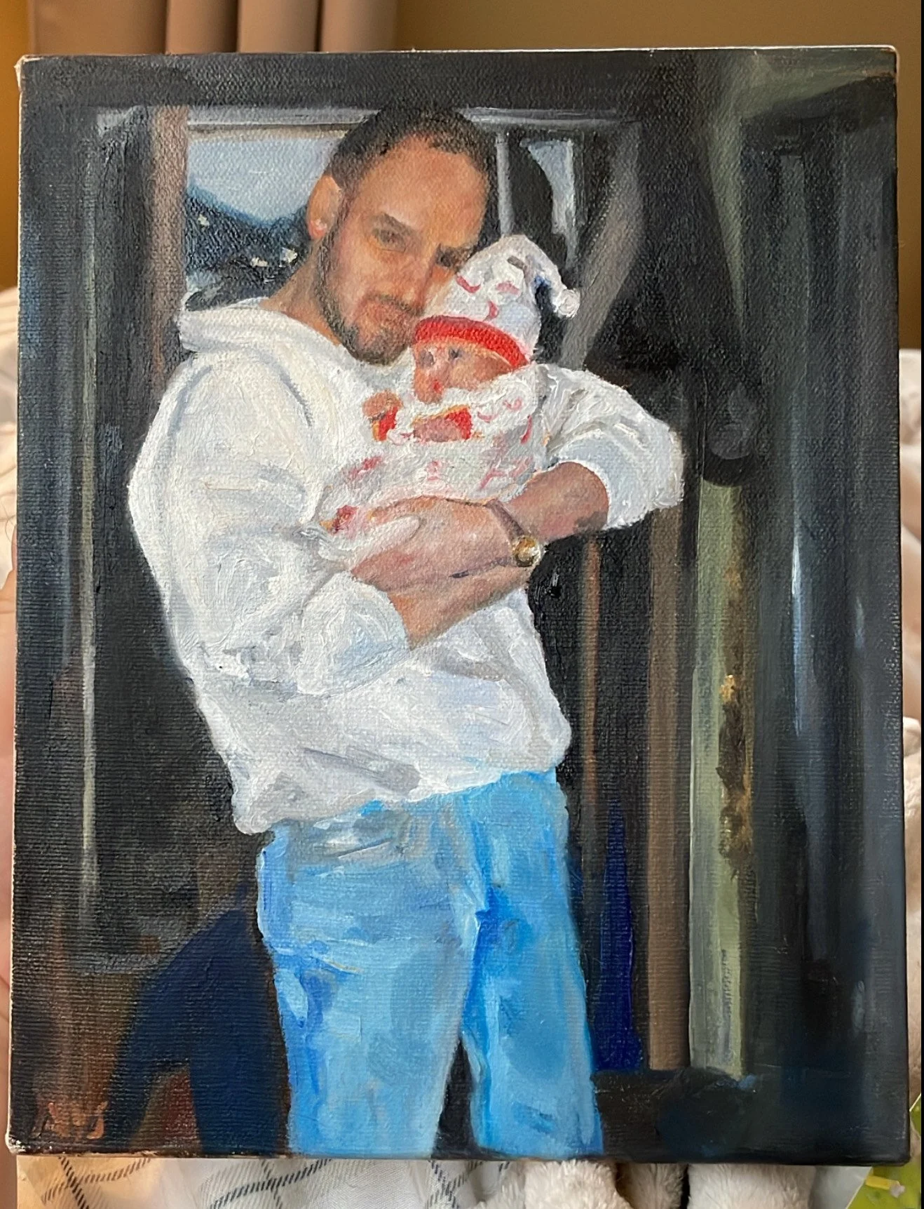 Painting of a man with a beard holding a baby in a white hoodie and colorful hat inside a room.