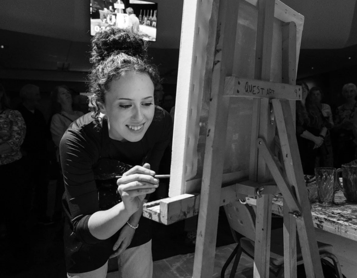A woman smiling and painting on a canvas set up on an easel at an event. There are people in the background watching.