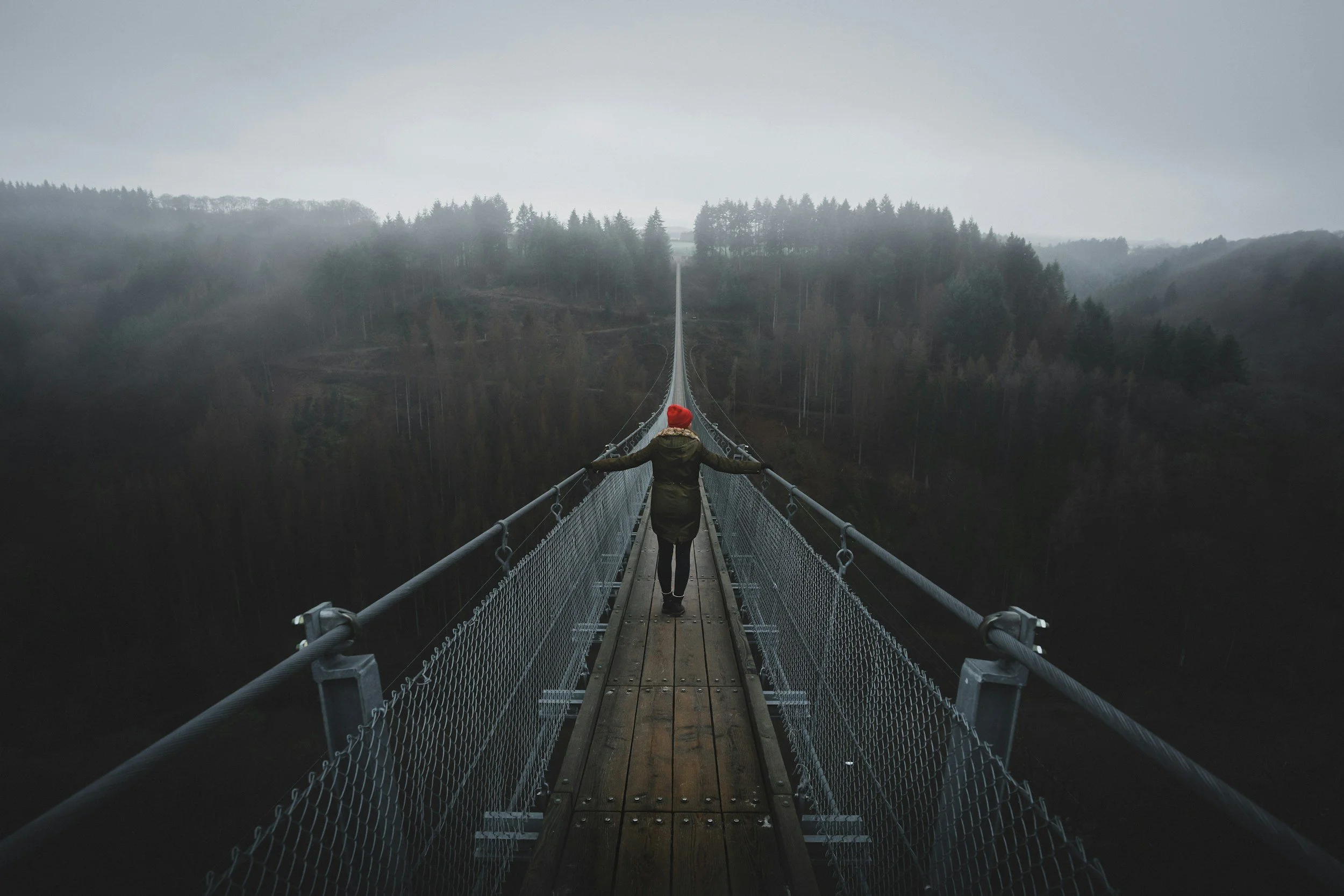 A woman walks across a long rope bridge into misty mountains, representing the journey from self-esteem to self-worth in negotiation and leadership.