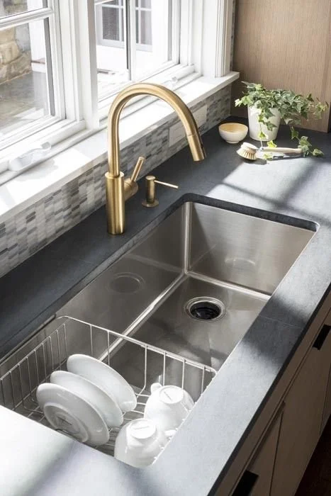 Kitchen sink with a gold faucet, dish rack with white dishes, window above, and small plants and bowls on the counter.