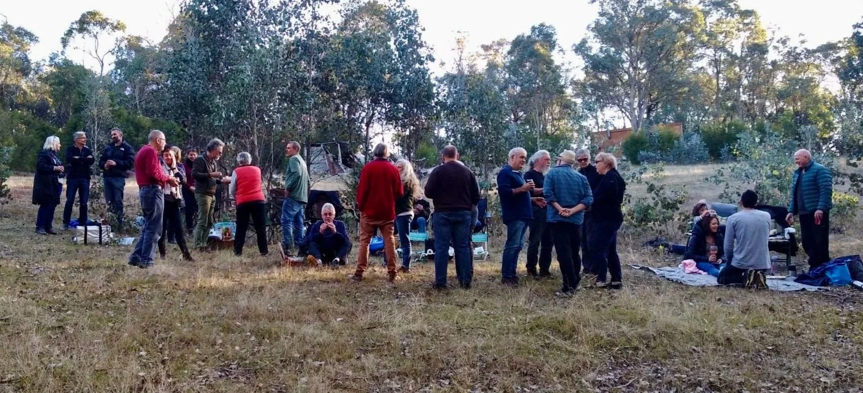 A group of people gathered outdoors in a natural setting with trees, some sitting on blankets or chairs, and others standing and chatting.