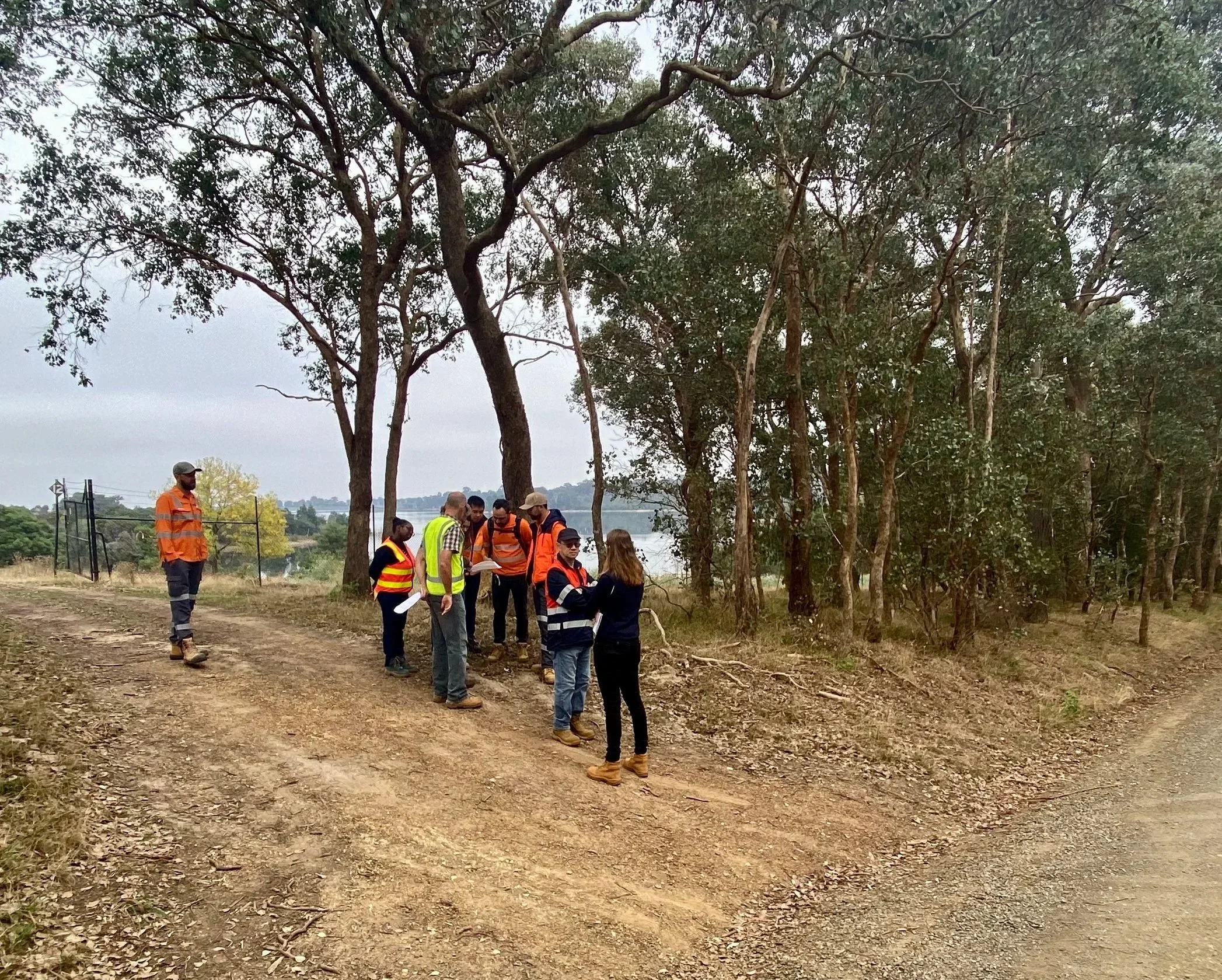 A group of people in safety vests and casual outdoor clothing standing on a dirt path next to trees, during overcast weather, possibly for a site inspection or outdoor work activity.