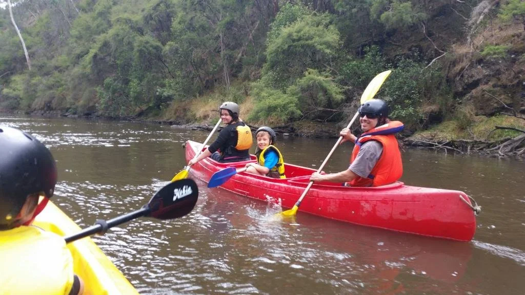 Family kayaking on river with two kids and one adult, surrounded by lush greenery.