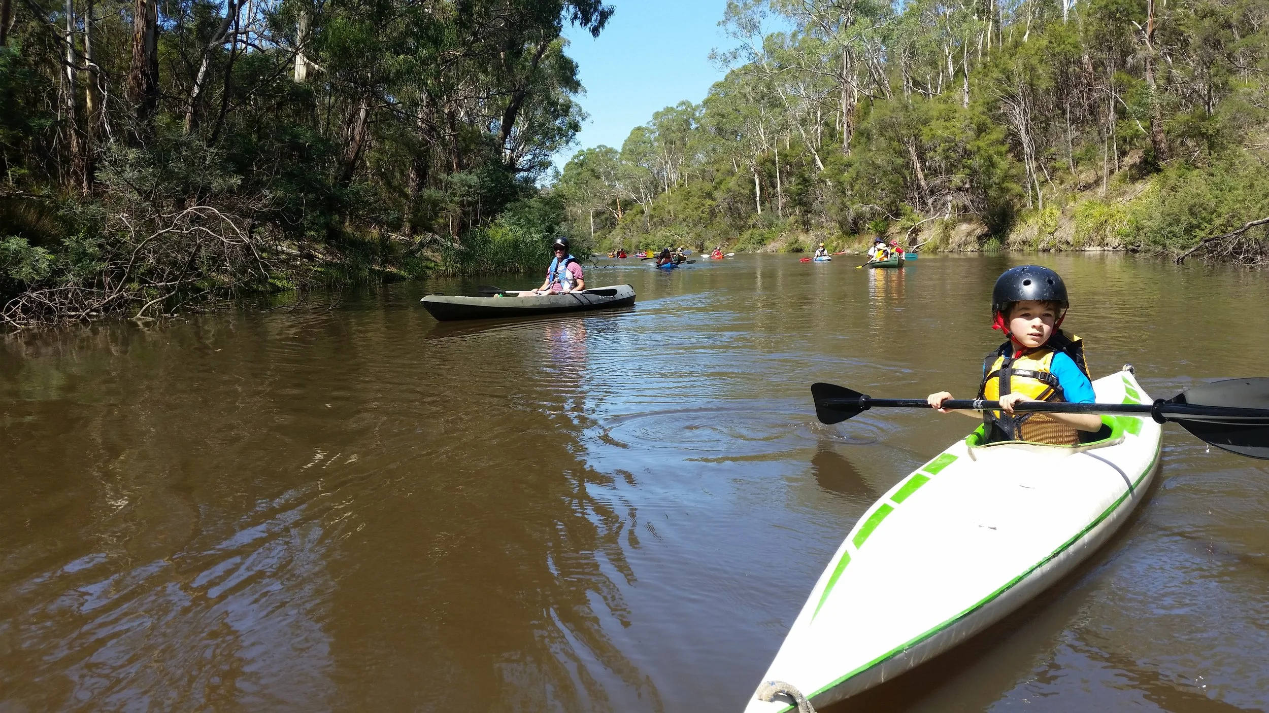 Young boy kayaking on a river, wearing a helmet and life vest, with other kayakers in the background amid trees and greenery.