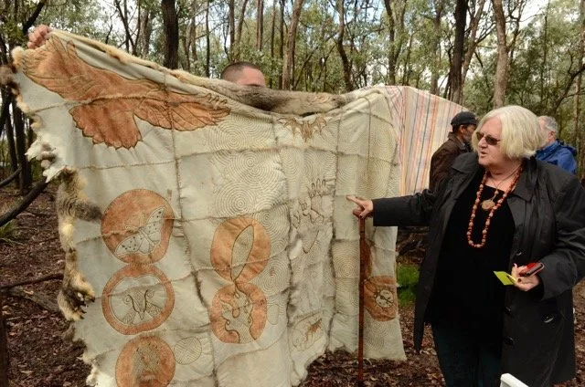 A woman with gray hair, sunglasses, a black coat, and a beaded necklace points at a large, decorated animal hide or textile hanging in a wooded outdoor setting, with a few other people nearby.