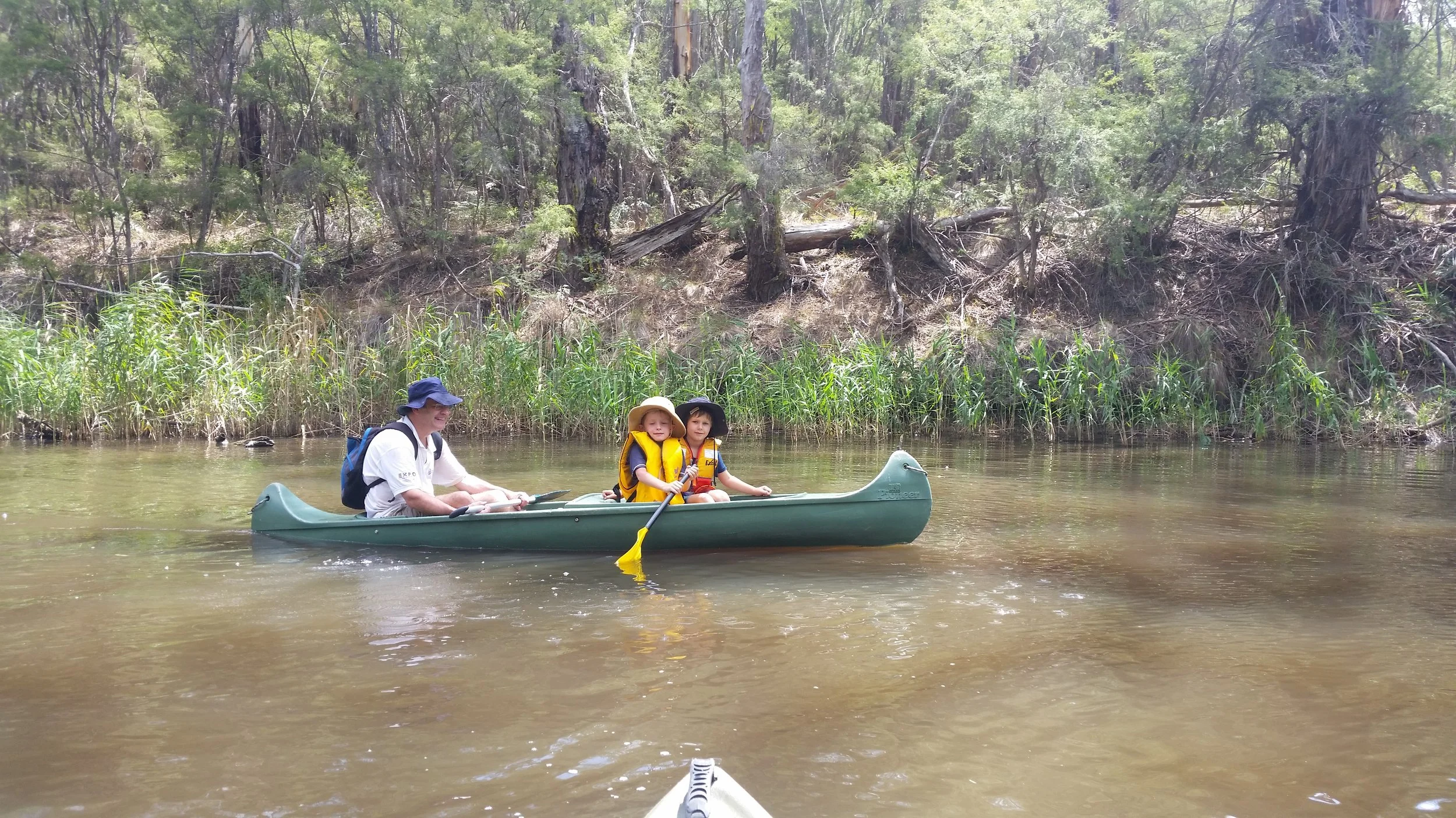 A man and two children in a green canoe on a river, surrounded by trees and greenery, with the man at the back and the children in the middle and front, paddling.