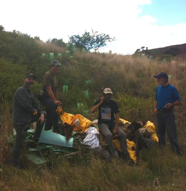 Four people in outdoor gear standing and sitting on a grassy hillside next to a crash site with debris and yellow pieces of equipment, under a partly cloudy sky.