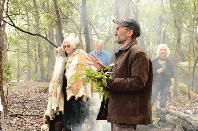 A man speaking outdoors in a wooded area with four women listening, some with glasses, one wearing a fur coat, others in jackets and sweaters, during the daytime.