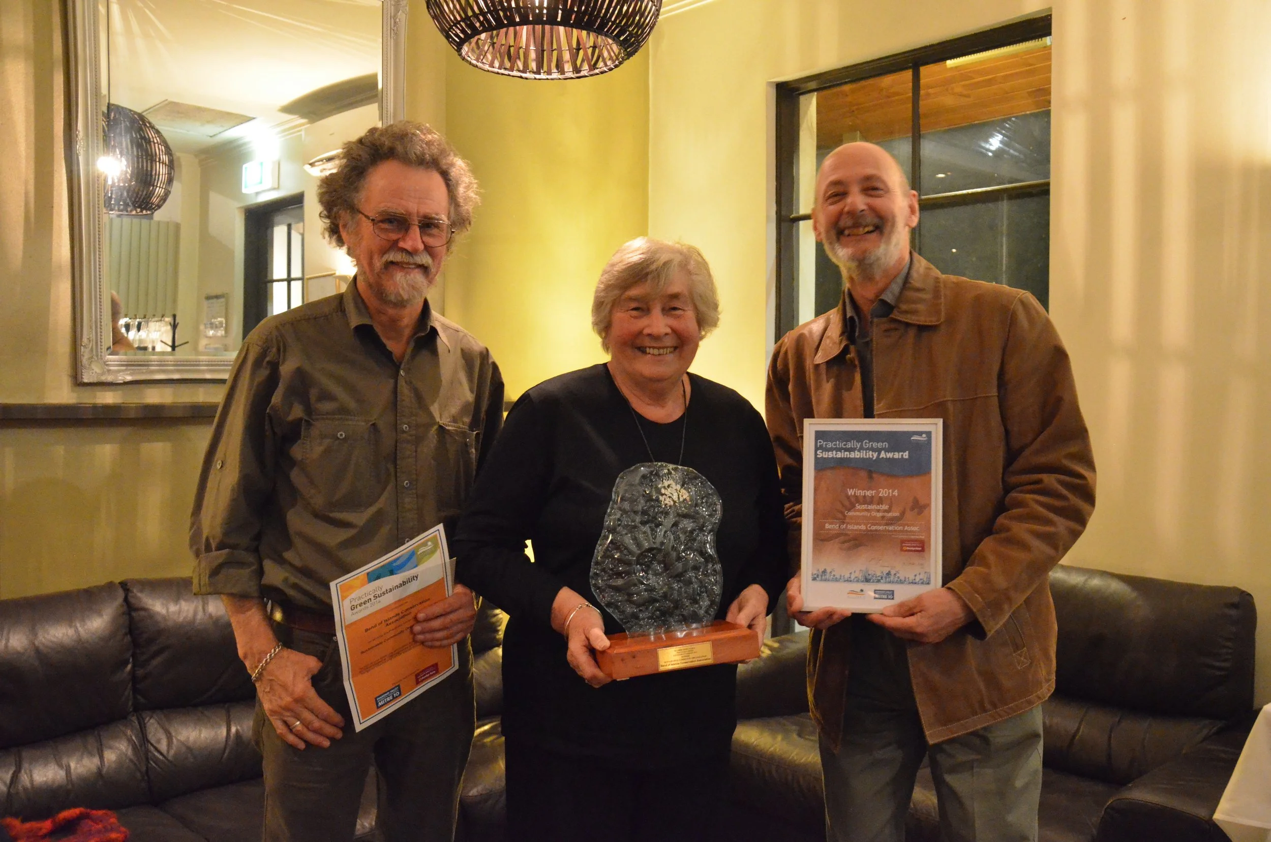 Three adults standing indoors, smiling. The woman in the middle holds a glass art award, and the man on the right holds a framed certificate. The man on the left also holds an orange and white certificate. The setting appears to be an awards ceremony or celebration in a warmly lit room with large windows and decorative lighting.
