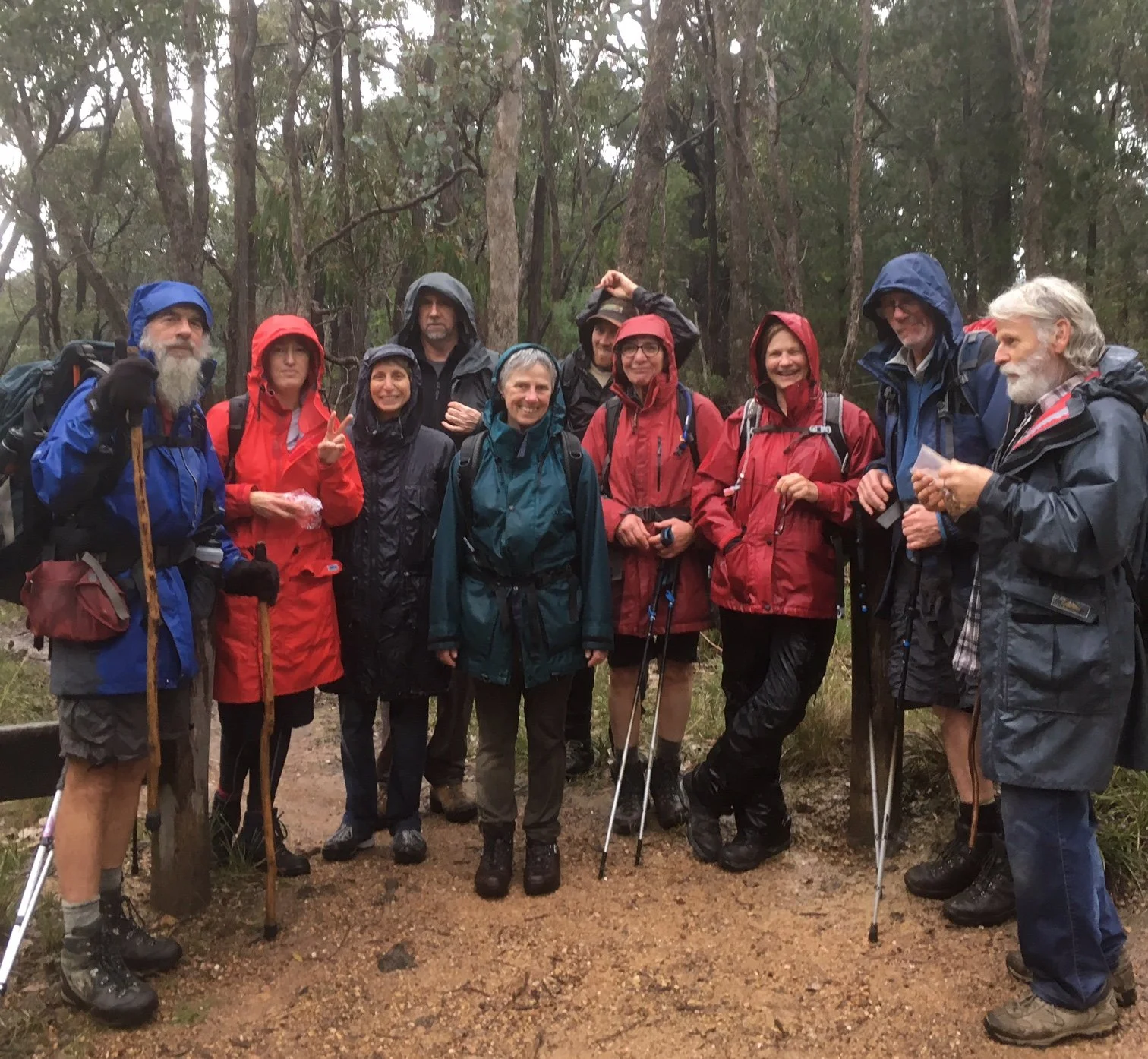 A group of people in outdoor rain gear and hiking boots standing on a trail in a forest, holding walking sticks and smiling.