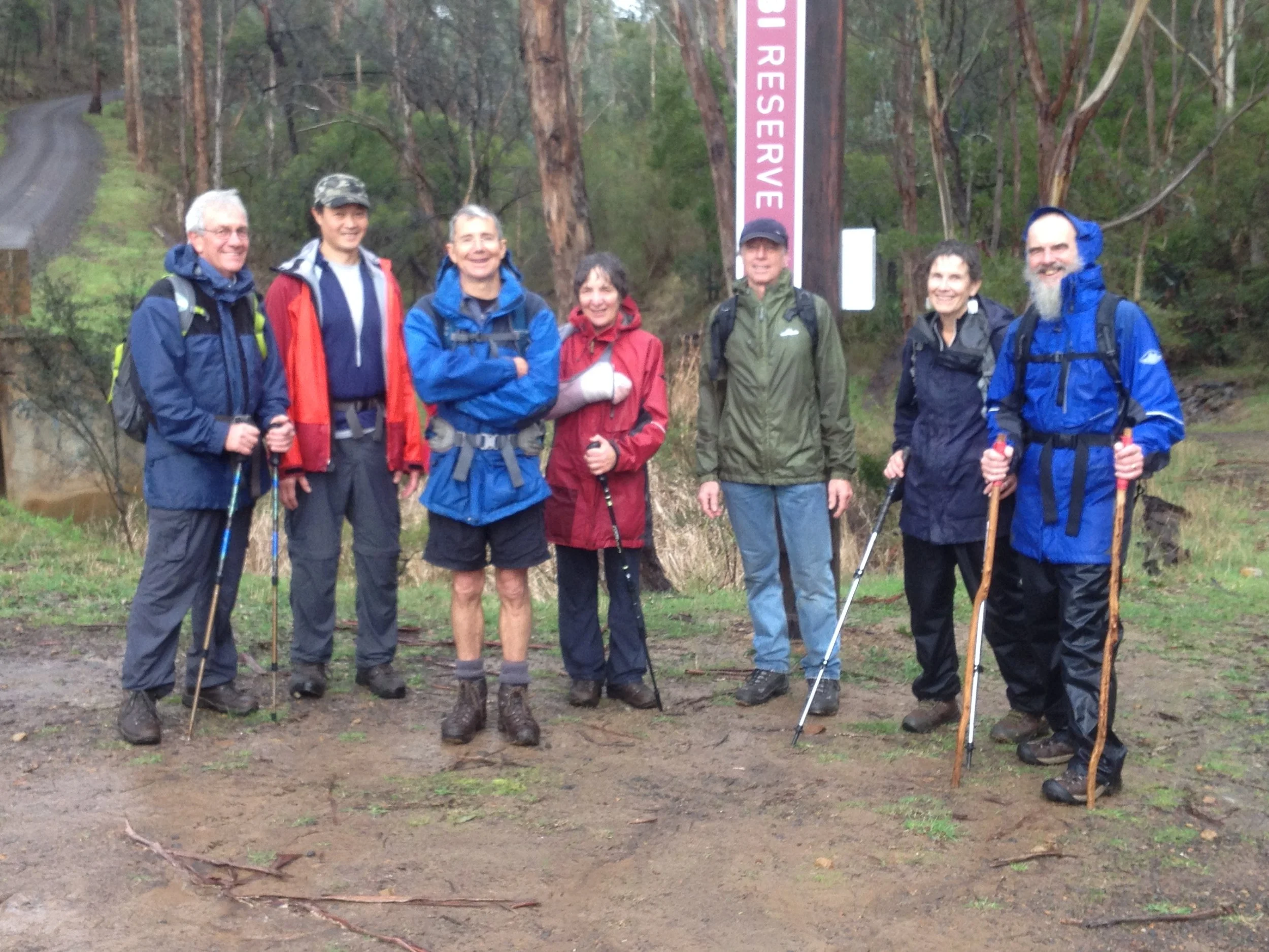 Group of seven people standing outdoors in hiking gear with backpacks, in a forested area near a sign that reads 'Resevoir'.