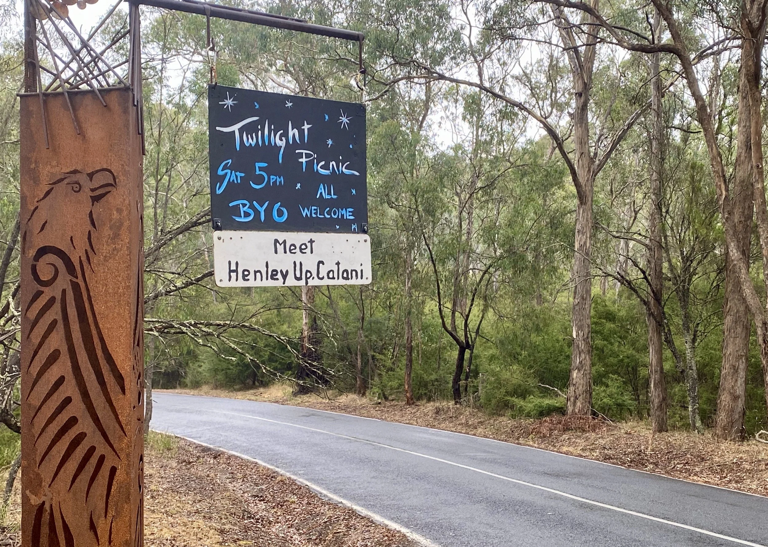 A sign hanging from a metal post along a wooded road, advertising a twilight picnic event on Saturday at 5 PM, open to all with BYO (bring your own) food, and inviting people to meet Henley Up Calani. The sign has a dark background with blue and white lettering and decorative stars, and there is a rust-colored metal post with an engraved bird and swirl design on it.