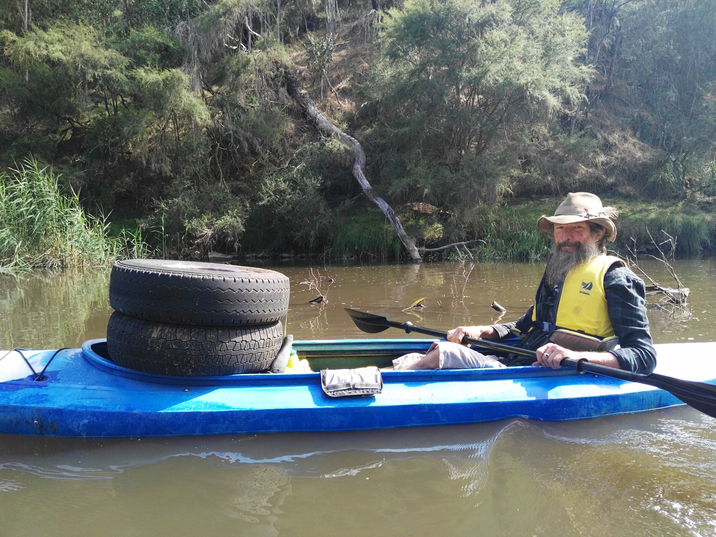 A man with a long beard, hat, and yellow life vest sitting in a blue kayak on a river, holding a paddle. Two tires are stored at the front of the kayak. The background features green vegetation and a fallen tree leaning over the water.