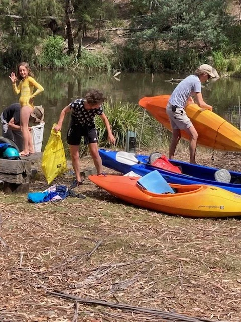 People preparing kayaks on the riverbank, with children and adults organizing gear near the water surrounded by trees.