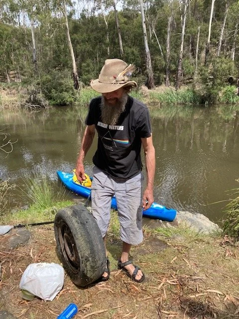 Man with a beard and a wide-brimmed hat standing near a water body, holding an old tire, with a blue kayak and some gear nearby in a forested area.