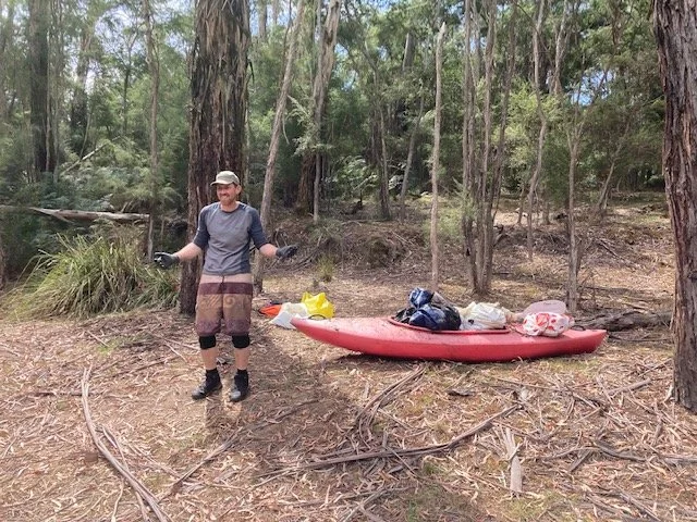 A man standing next to a red kayak on a forested lakeside, surrounded by trees and natural scenery, with gear and supplies on the kayak.