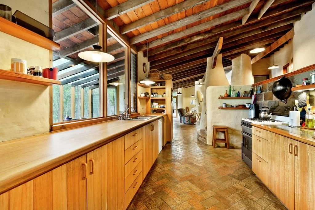 A rustic kitchen with wooden cabinets, a brick floor, exposed wooden ceiling beams, and large windows overlooking greenery.