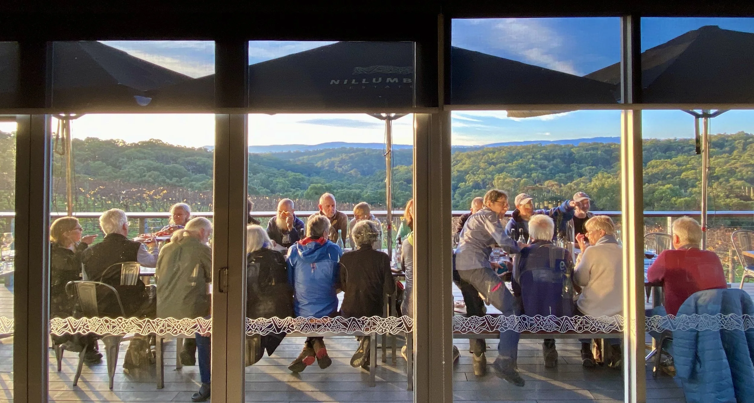 Group of people dining outdoors on a balcony with a scenic view of green hills and blue sky in the background, seen through large glass windows.