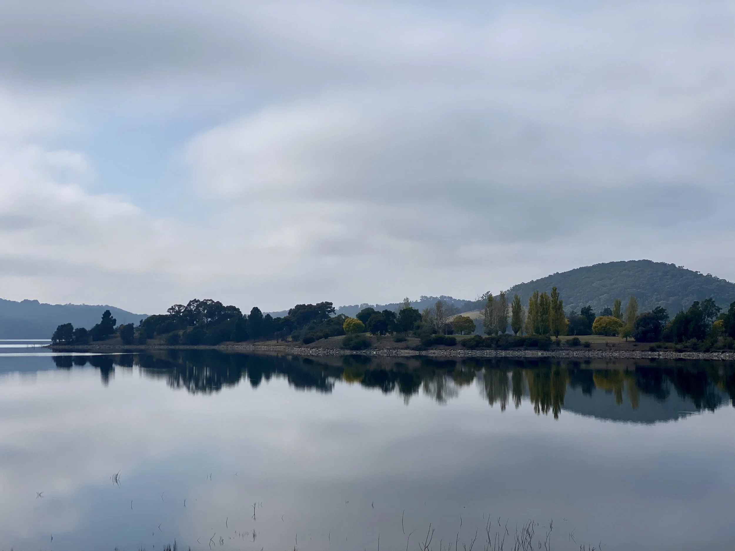 Scenic view of a calm lake with a reflection of trees and hills under a cloudy sky.