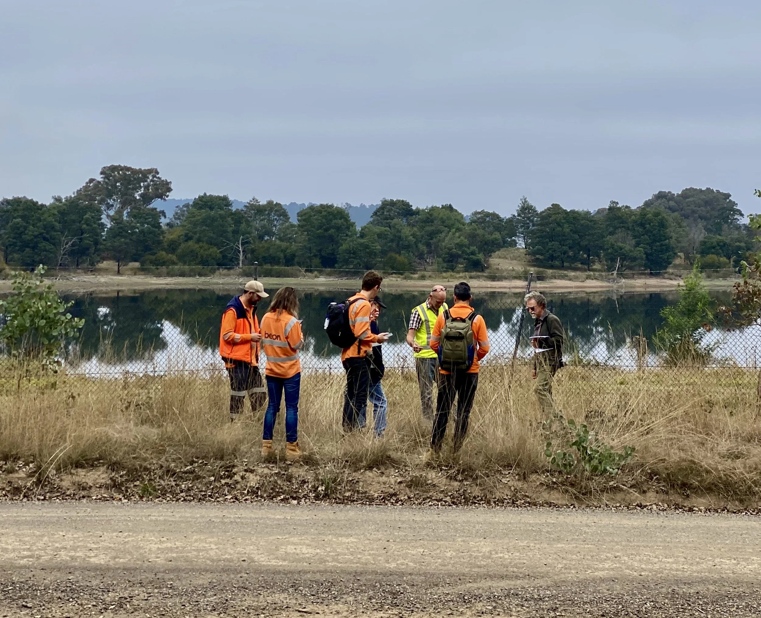 Group of six people, some wearing orange safety vests, standing near a fence by a body of water, with trees and overcast sky in the background.