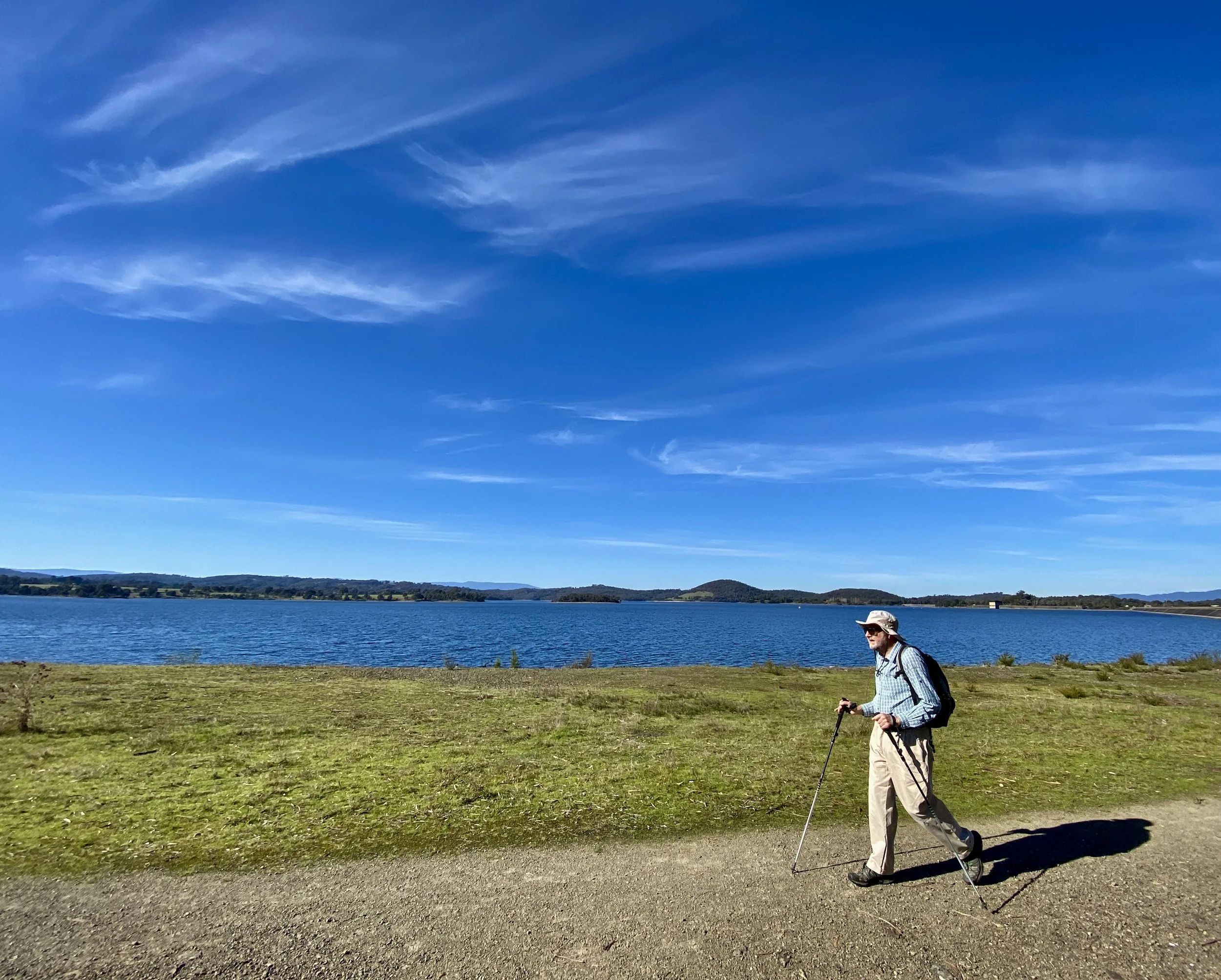 An elderly man hiking with a walking stick along a dirt path beside a large body of water, with hills and a clear blue sky in the background.