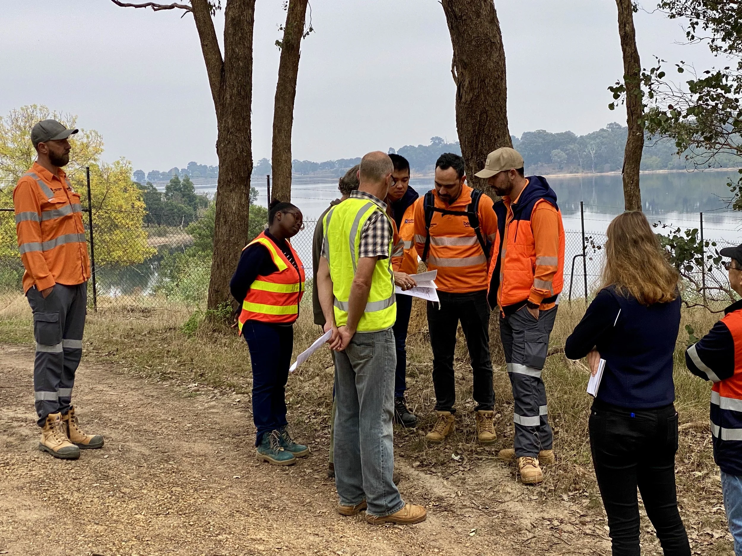 A group of people, some wearing high-visibility safety vests, standing outdoors near trees and a body of water, gathered around a man who is showing them documents.