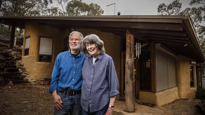 An older man and woman standing outside a rustic house with stucco walls and wooden accents, both smiling.