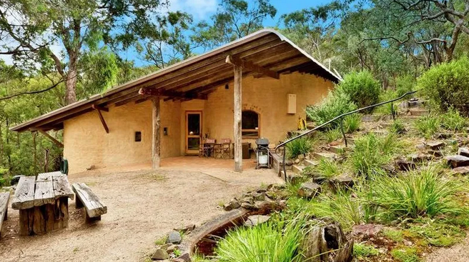 A rustic house surrounded by trees with a front porch, outdoor seating, and a sloped pathway with rocks and greenery.