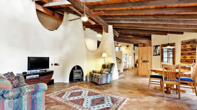 Living room with a colorful patterned sofa, an armchair, a television, a fireplace, and a rug. Dining area with a wooden table and chairs in the background, with exposed wooden beams on the ceiling and a rustic style.