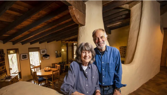 An elderly woman and man smiling in a rustic home interior with wooden beams and art on the walls.
