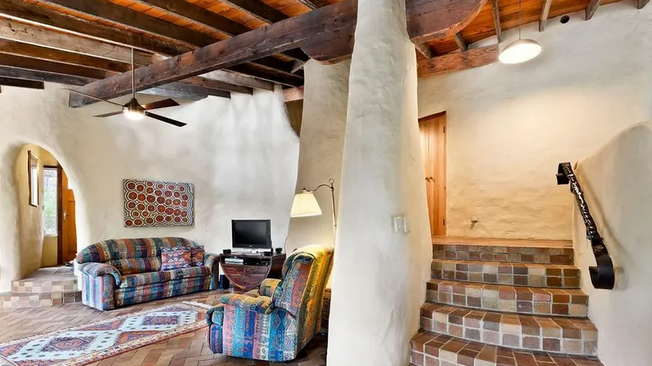 Living room with rustic wood ceiling, stucco walls, brick staircase, upholstered furniture, ceiling fan, and a flat-screen TV.