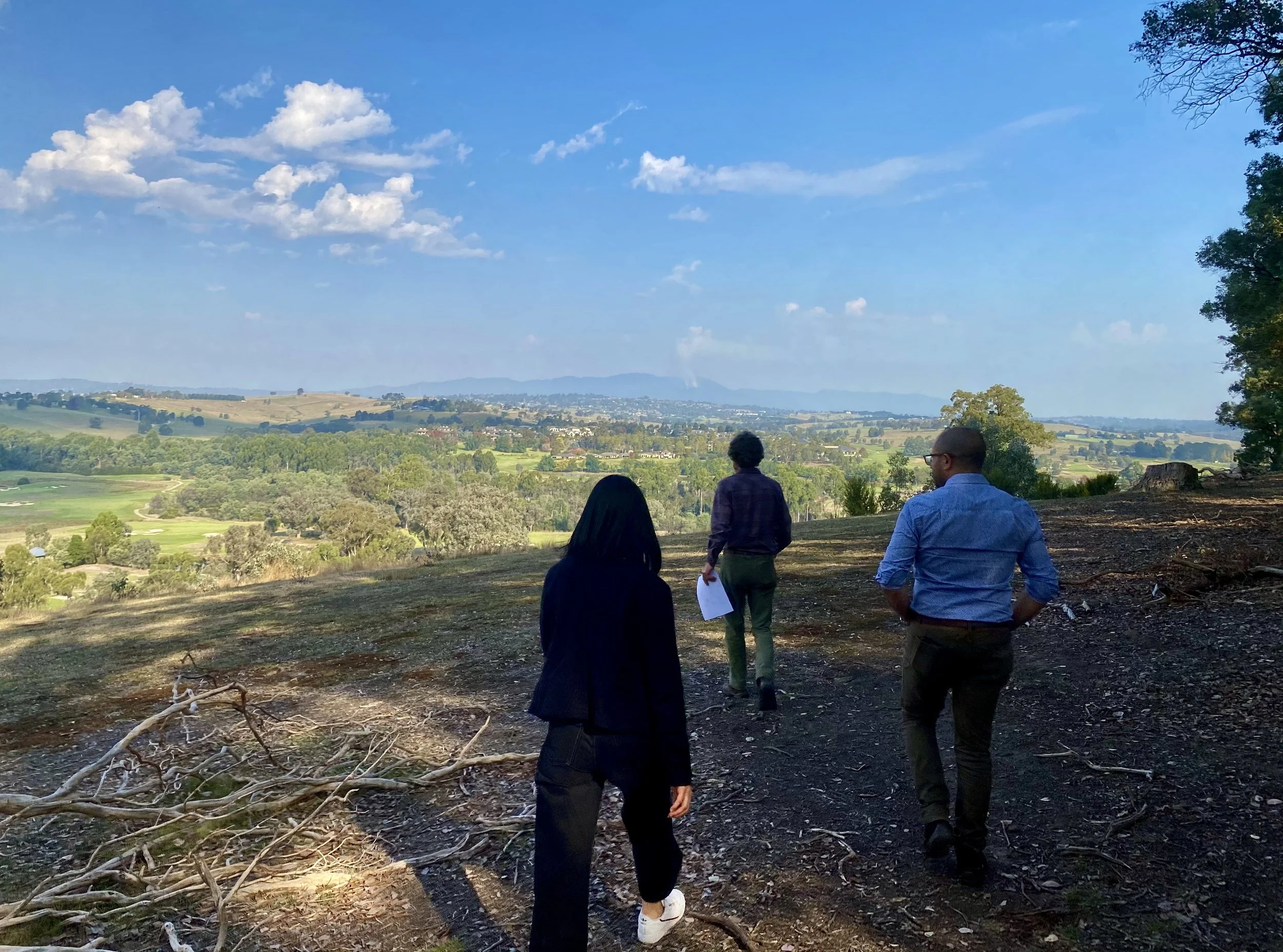 Three people walking on a dirt path overlooking a vast green landscape with rolling hills, trees, and a blue sky with clouds.